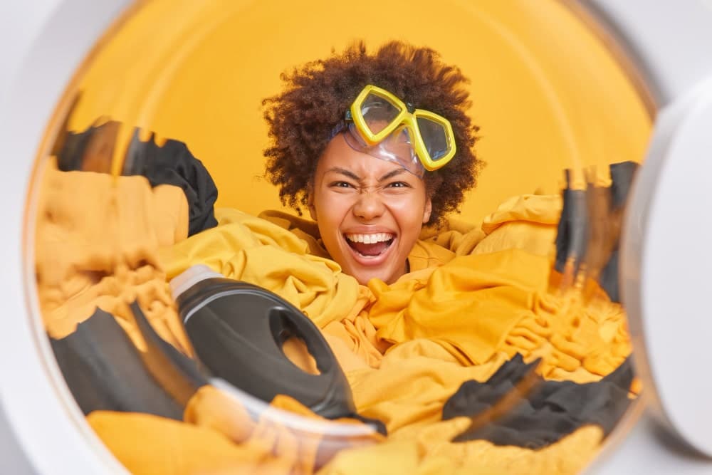funny housewife with curly hair wears snorkeling mask forehead poses inside washing machine surrounded by yellow black dirty clothes puts laundry washer min